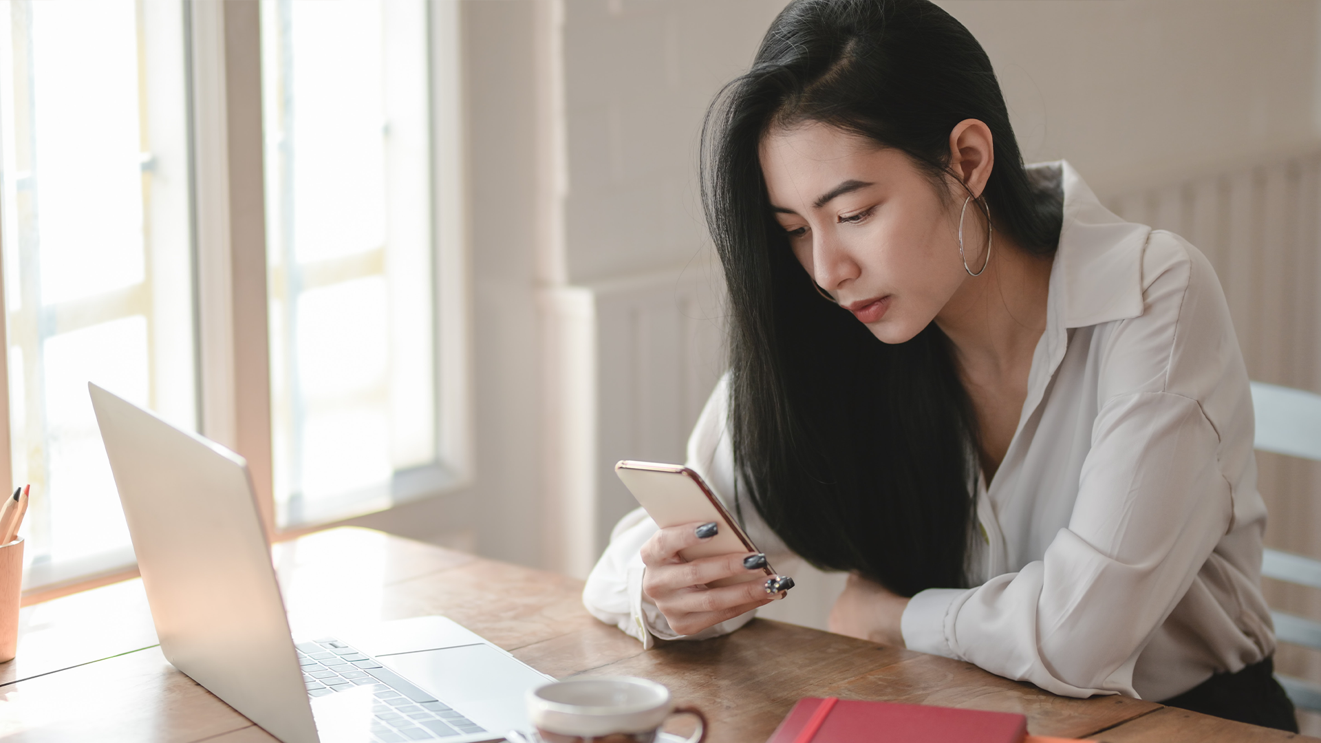 A woman sitting at a table with a laptop in front of her. She is looking at her smartphone.