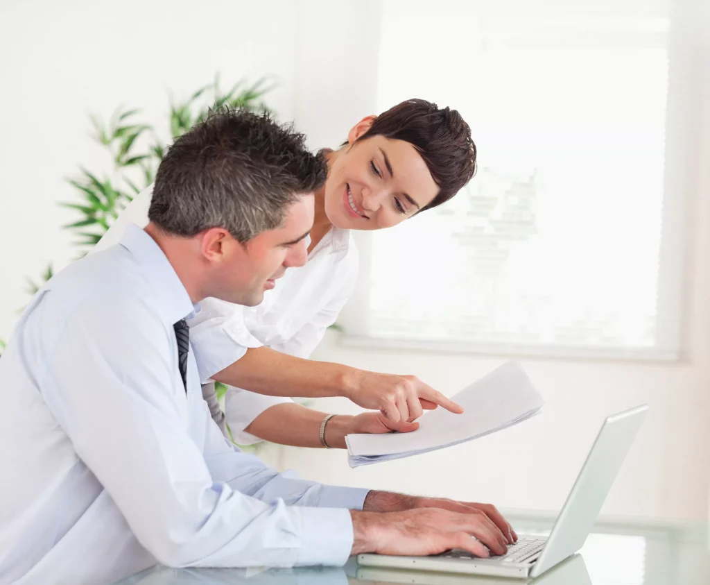 A woman pointing to a piece of paper and a man looking at the paper while working on a laptop.