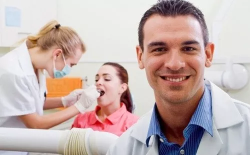 Dentist in foreground smiling at camera and dental assistant in background working with a patient.