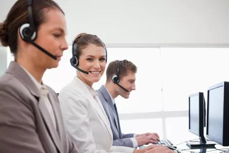 People wearing headsets sitting at computers, working. One person sitting in the middle is smiling at the camera.