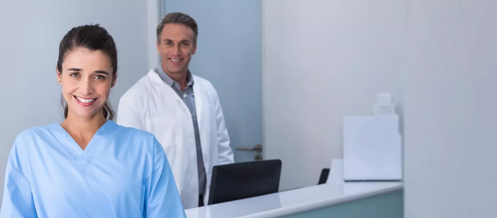 Two dental professionals in a dental office setting smiling at the camera. A computer is beside them on the right.