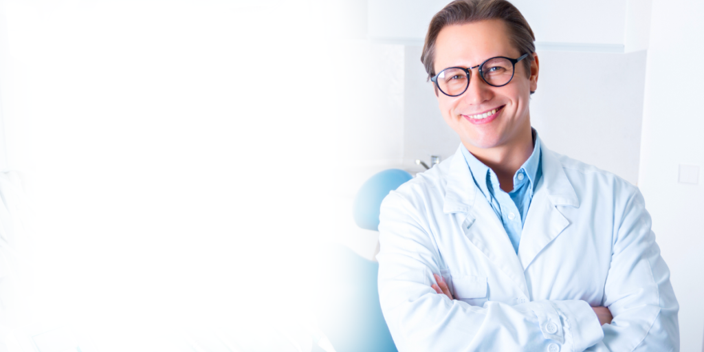 A male dentist smiling and looking at the camera with crossed arms. The dentist is wearing a lab coat and glasses.